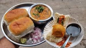 "Indian street food combo of vada pav and pav bhaji served on a plate with chopped onions, lemon wedge, and buttered pav